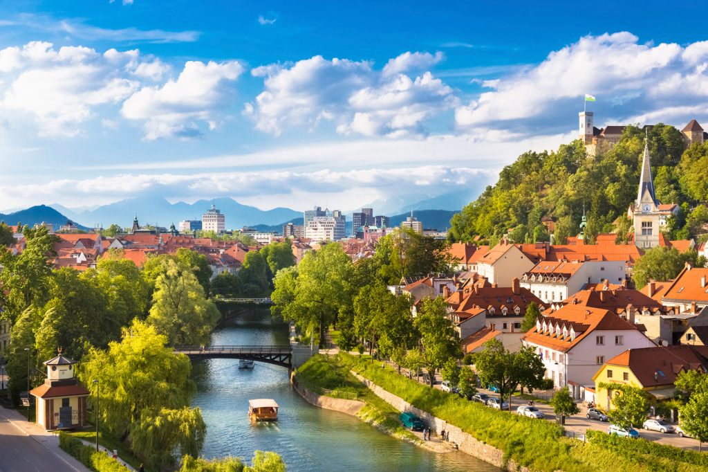 Ljubljana, Slovenia landscape, with the river running through the middle of the image. Buildings are on either side, with lush trees filling the remainder of the space.