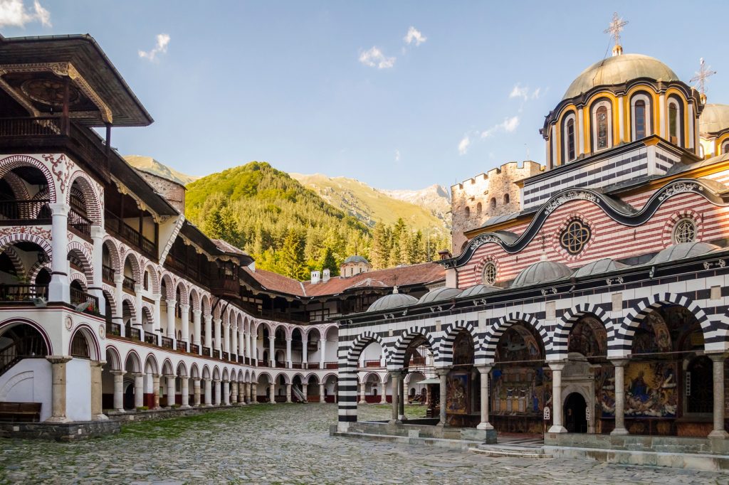 Sofia, Bulgaria is a budget-friendly destinations in Europe. Mountainous landscape in the background, with a church-like building in the forefront. The building has multiple arches.
