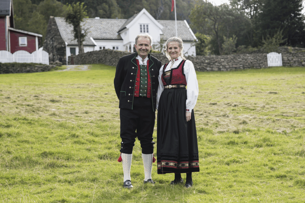 A man and woman dressed in traditional norwegian clothing stand in front of a house.