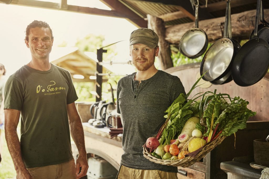 Two men standing in front of a kitchen with a basket of vegetables.