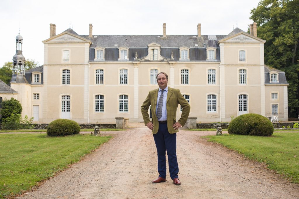 A man in a suit standing in front of a large mansion.
