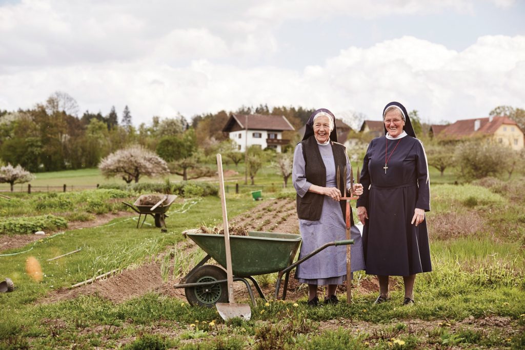 Two nuns standing next to a wheelbarrow in a garden.