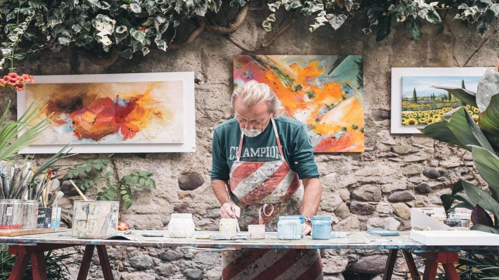 A man in an apron paints at a table, enjoying his second career in front of a garden.