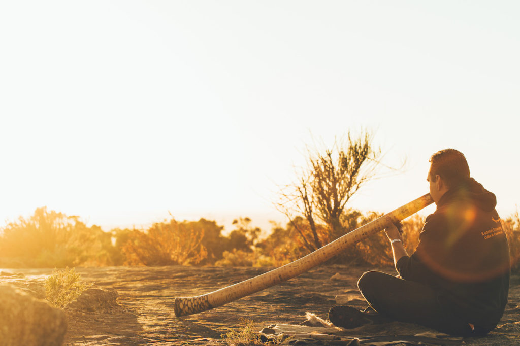 A man sitting on the ground with a wooden stick in his hand, experiencing an Australian adventure.