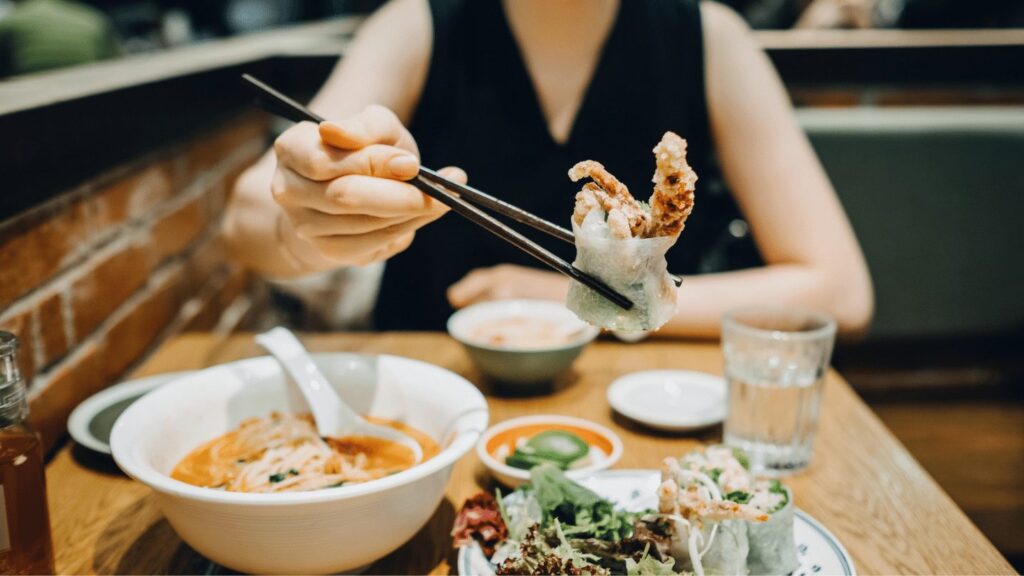 A woman using chop sticks to each a variety of Vietnamese foods.