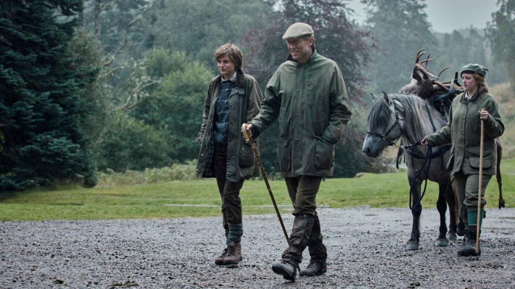 Three individuals walking outdoors with a reindeer and a pony on a gravel path, wearing wet weather attire, reminiscent of scenes from "The Crown Season 4.