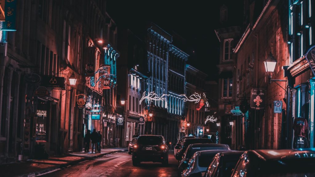 A nighttime city street illuminated by streetlights and adorned with Christmassy decorations, with cars and pedestrians visible.