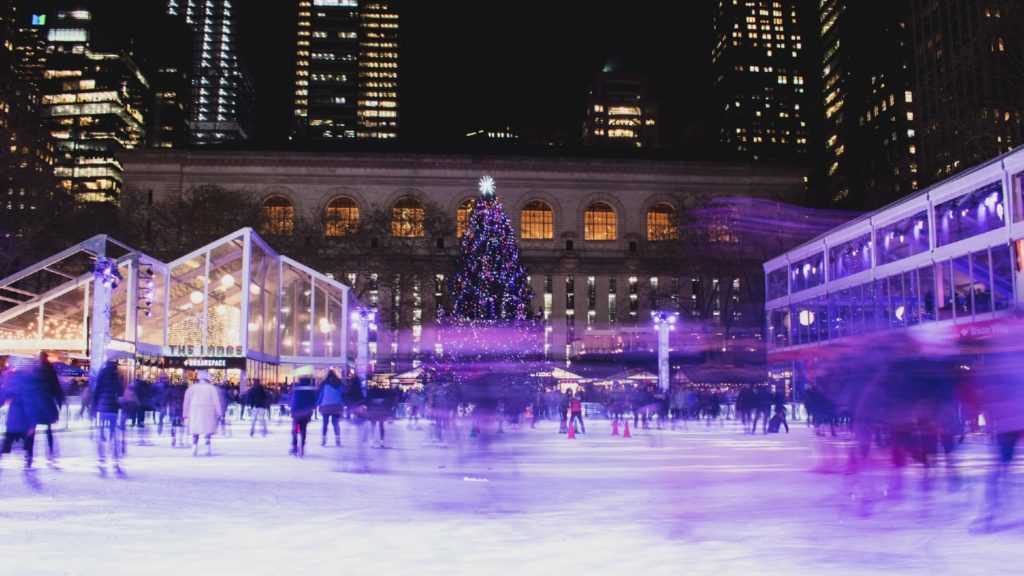 Ice skaters glide across a rink with a large Christmas tree illuminating the evening, Christmassy places to visit and city buildings in the background.