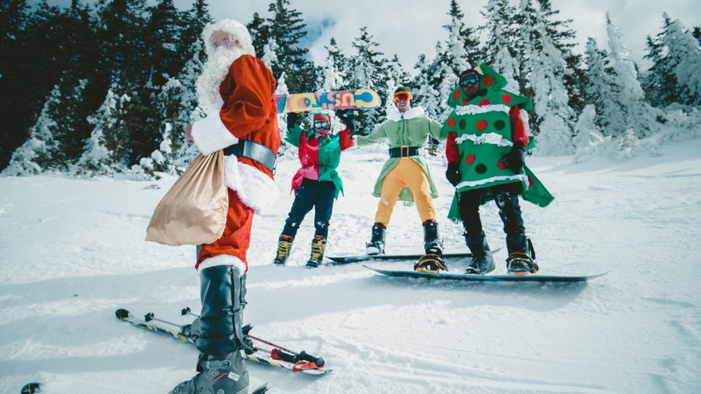 Individuals dressed in Santa Claus and Christmas tree costumes with a snowboarder on a snowy mountain explore Christmassy places to visit.