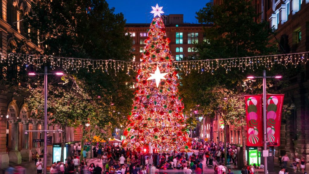 A festive crowd enjoys a brightly lit Christmas tree at a city evening event in Sydney.