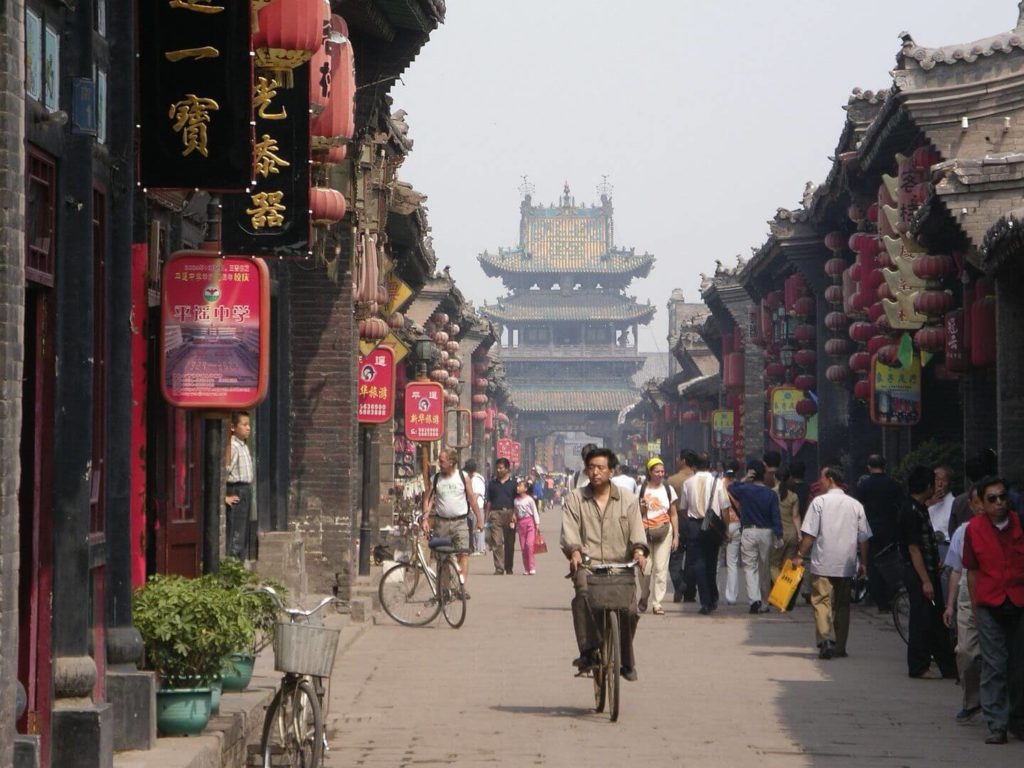 A bustling street scene in a historic Chinese town with traditional architecture and pedestrians offers a glimpse into the "things to know before traveling to China.