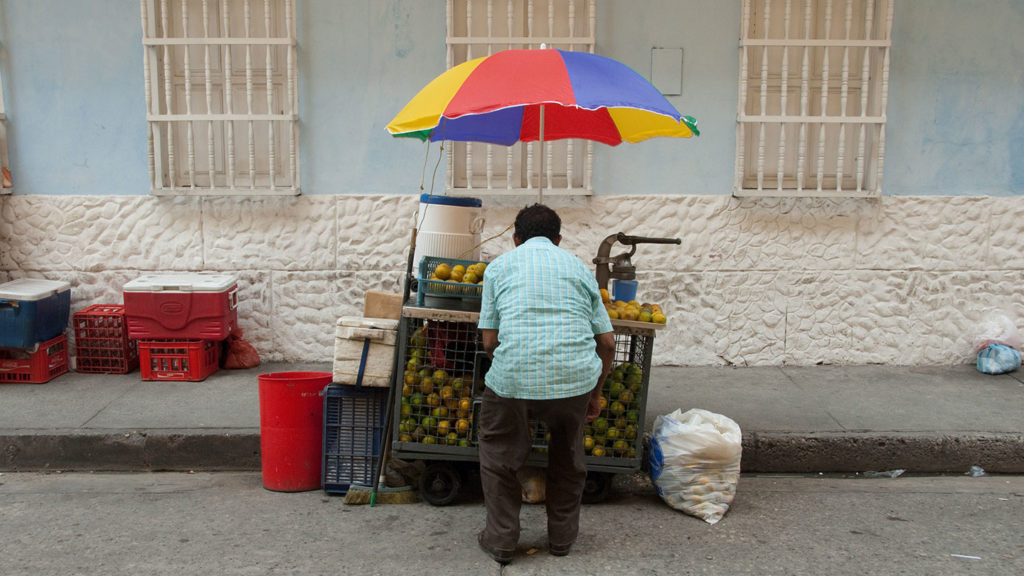 A vendor stands at a fruit cart under a colorful umbrella on a city street, catering to tourists on the gringo trail in South America.
