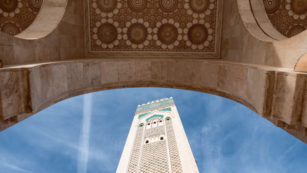 A view of an ornately decorated minaret through an arch with intricate mosaic patterns under a clear blue sky, perfect for a wedding anniversary travel destination.