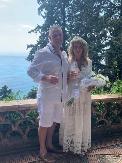 A couple dressed in white holding glasses and flowers stands together on a balcony with a view of the sea in the background during their Trafalgar trip.