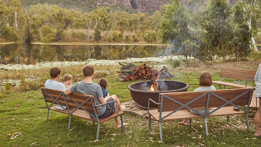 A group of people relaxing by a campfire near a waterbody with lily pads, surrounded by the natural landscape during their tour of Outback Australia.