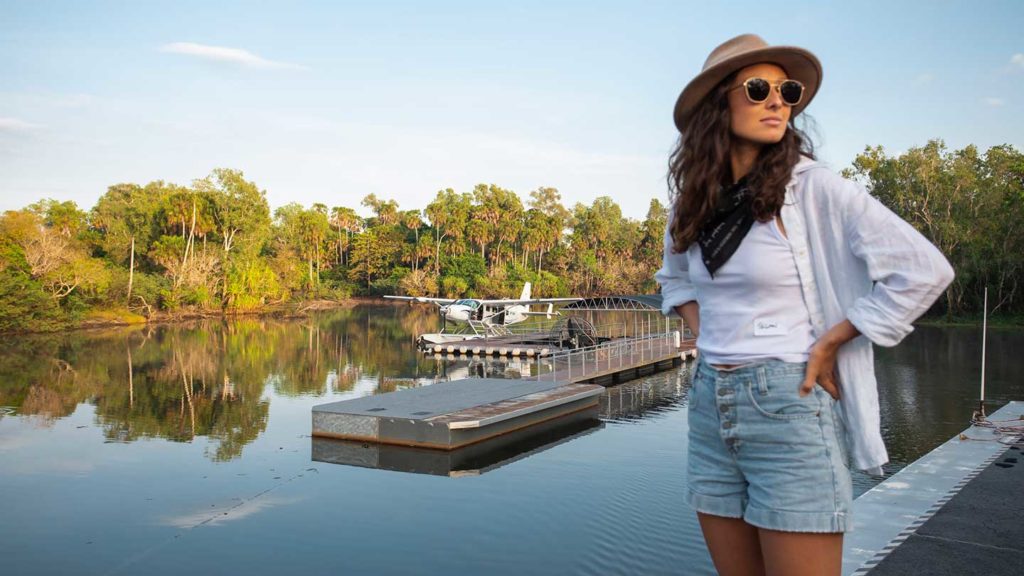 Woman in summer attire standing by a river dock with a small boat in the background, enjoying her outback Australia trip.