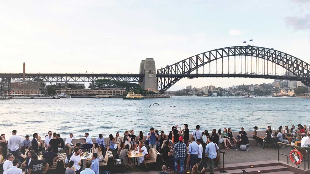 Group of people enjoying an outdoor gathering near a waterfront with the Sydney Harbour Bridge in the background, contemplating what to pack for their next east coast Australia travel adventure.