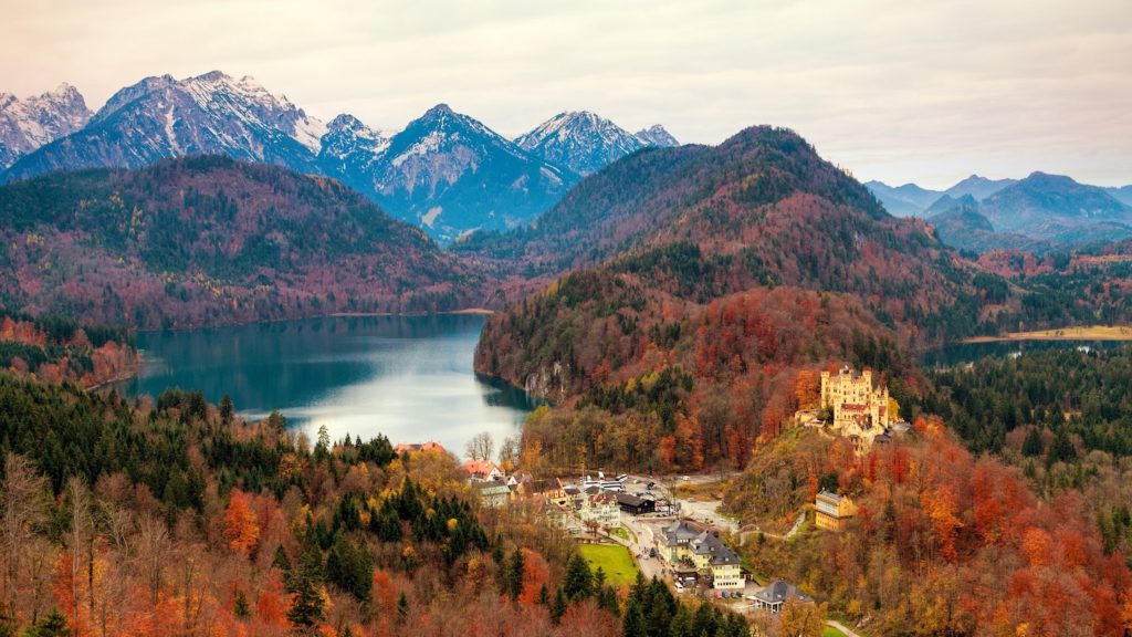 Autumn panorama of one of the most beautiful Bavarian castles nestled in the foothills with a backdrop of snow-capped mountains and a tranquil lake.