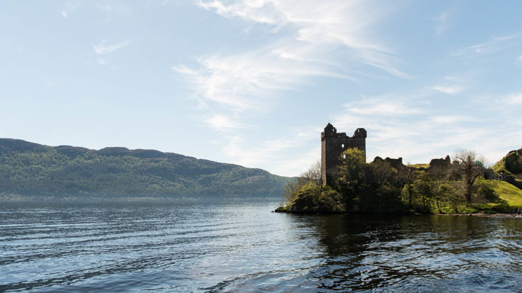 Ruined castle on a lakeshore with hills in the background in Scotland on a sunny day.