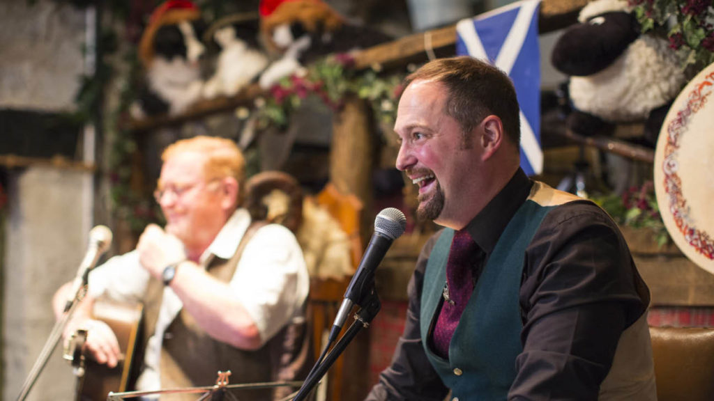 Two men in traditional Scottish attire performing at a festive event with decorations and musical instruments.