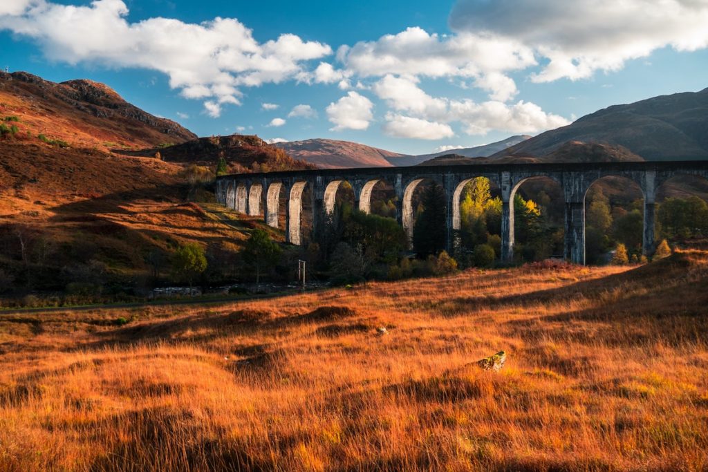 Stone viaduct amid autumnal landscape with rolling hills and orange-hued grasses under a partly cloudy sky, one of the UK's best film locations.
