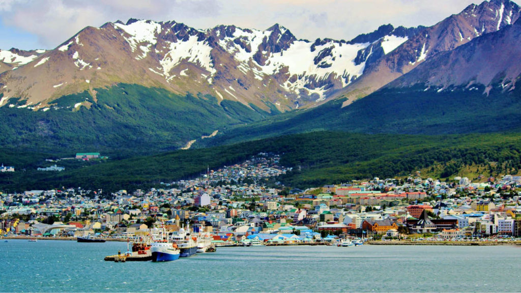A vibrant coastal town nestled at the foot of a mountain range in Patagonia, with ships docked in the foreground.