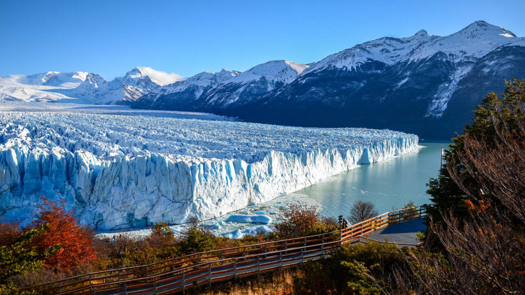 Glacier extending into a UNESCO-listed lake with surrounding mountains and a clear blue sky, viewed from a wooden walkway.