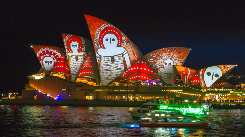 Sydney Opera House illuminated with red and white Aboriginal-inspired patterns at night, with a boat passing by, becomes one of the best things to do in Sydney.