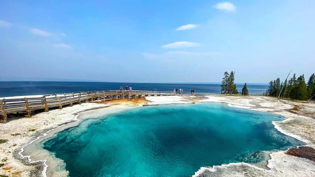 A wooden boardwalk encircles a vibrant blue hot spring, a must-see lake in the US, with visitors exploring the area against a backdrop of a forest and hazy sky.