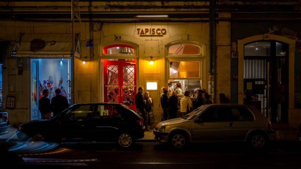 A group of people gathering outside a warmly-lit restaurant named "tapisco" at night, celebrating quarantine-free travel, with cars parked in front.