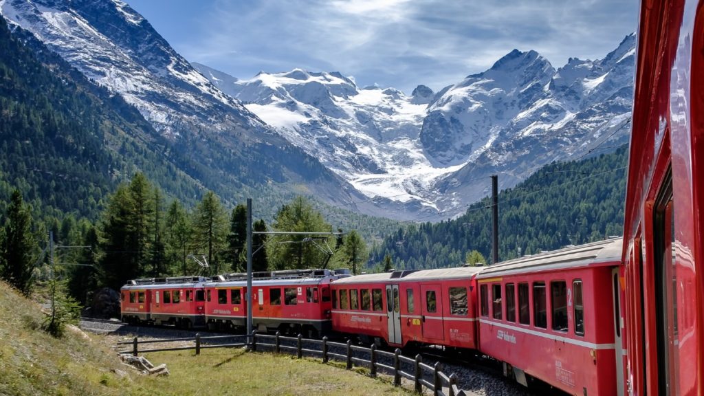 Red passenger train traveling through a mountainous landscape with snow-capped peaks in the background, offering one of the world's best train journeys.
