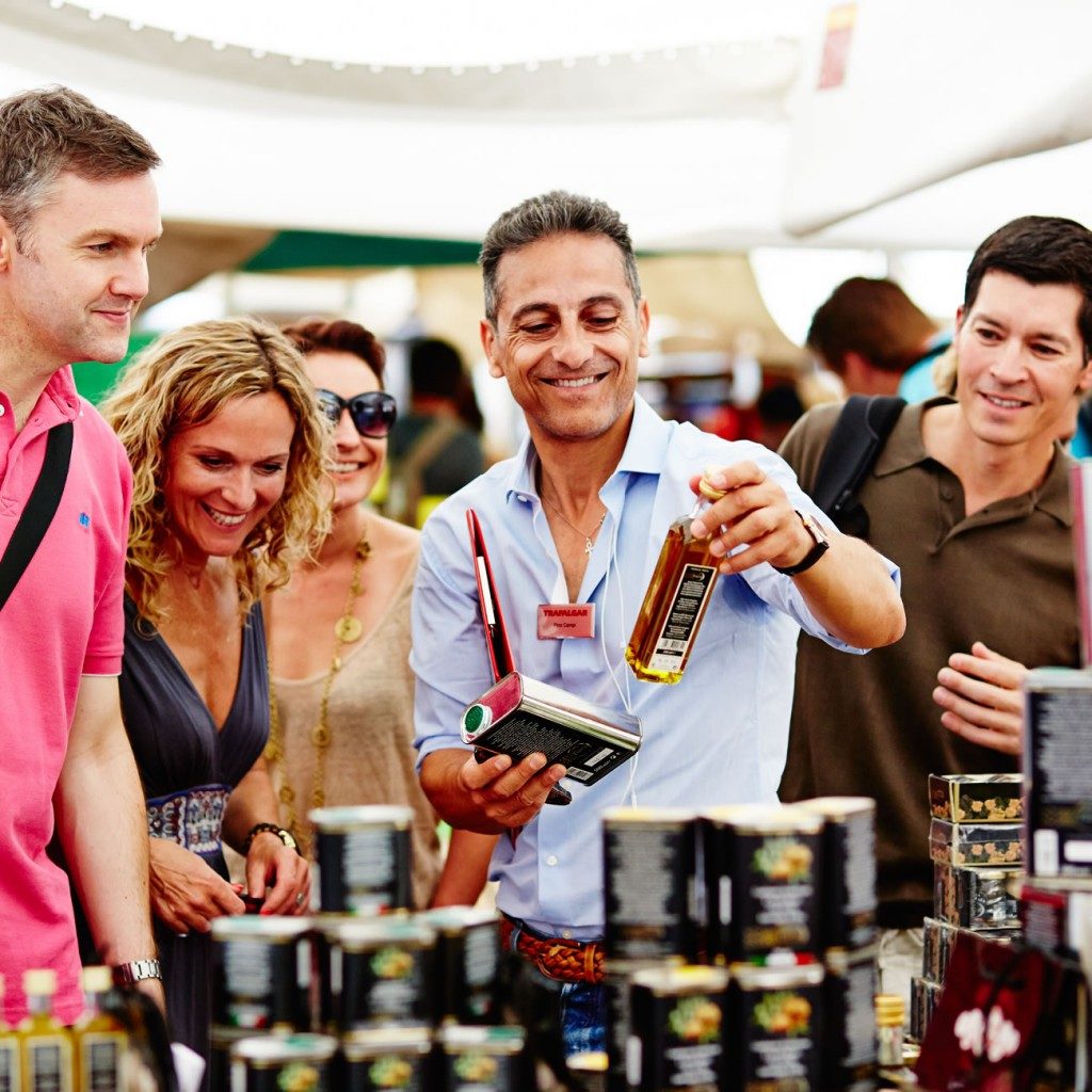 A vendor offers a sample to smiling customers at a European food market stall.