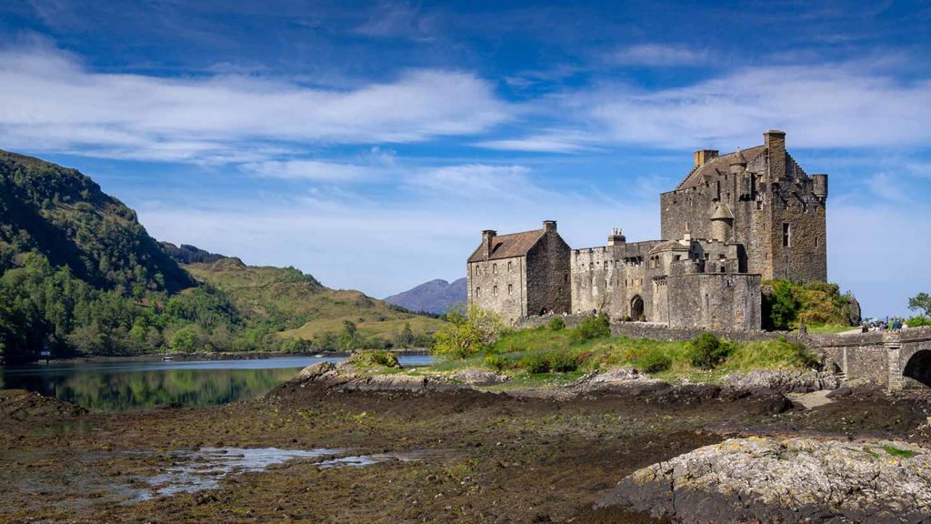 Historic castle beside a calm lake with surrounding greenery under a blue sky, known as one of the James Bond filming locations.