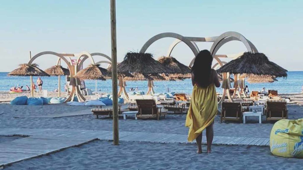 Woman in a yellow cover-up standing on the beach, looking out at the sea with straw umbrellas and sun loungers in the foreground, capturing the essence of summer in Switzerland.