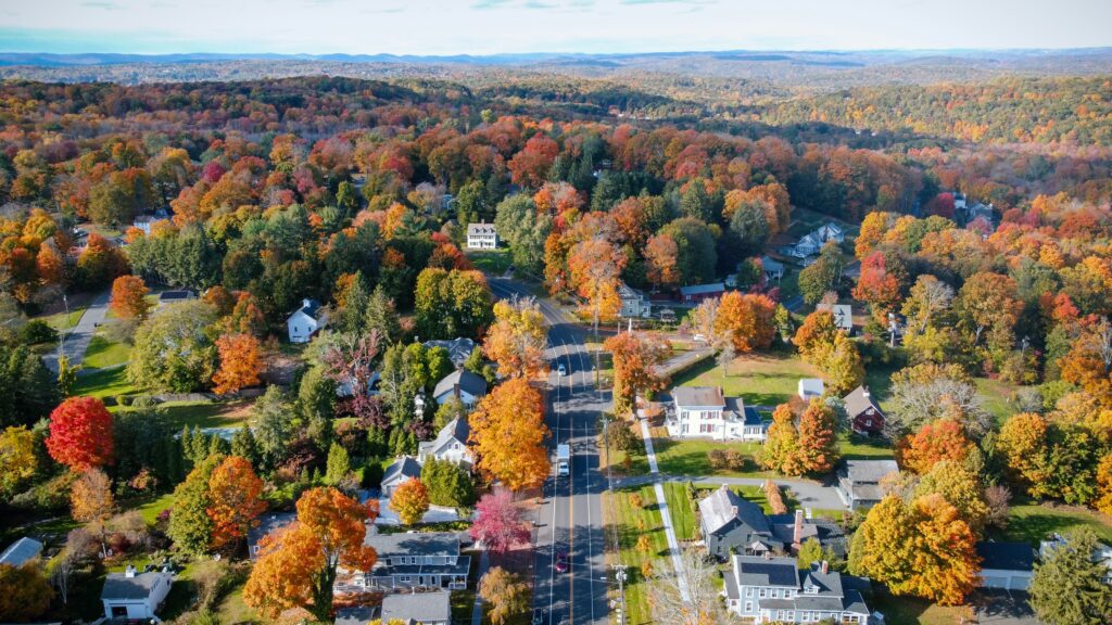 A quaint New England town road bisecting forests of orange, yellow, and brown autumn trees.