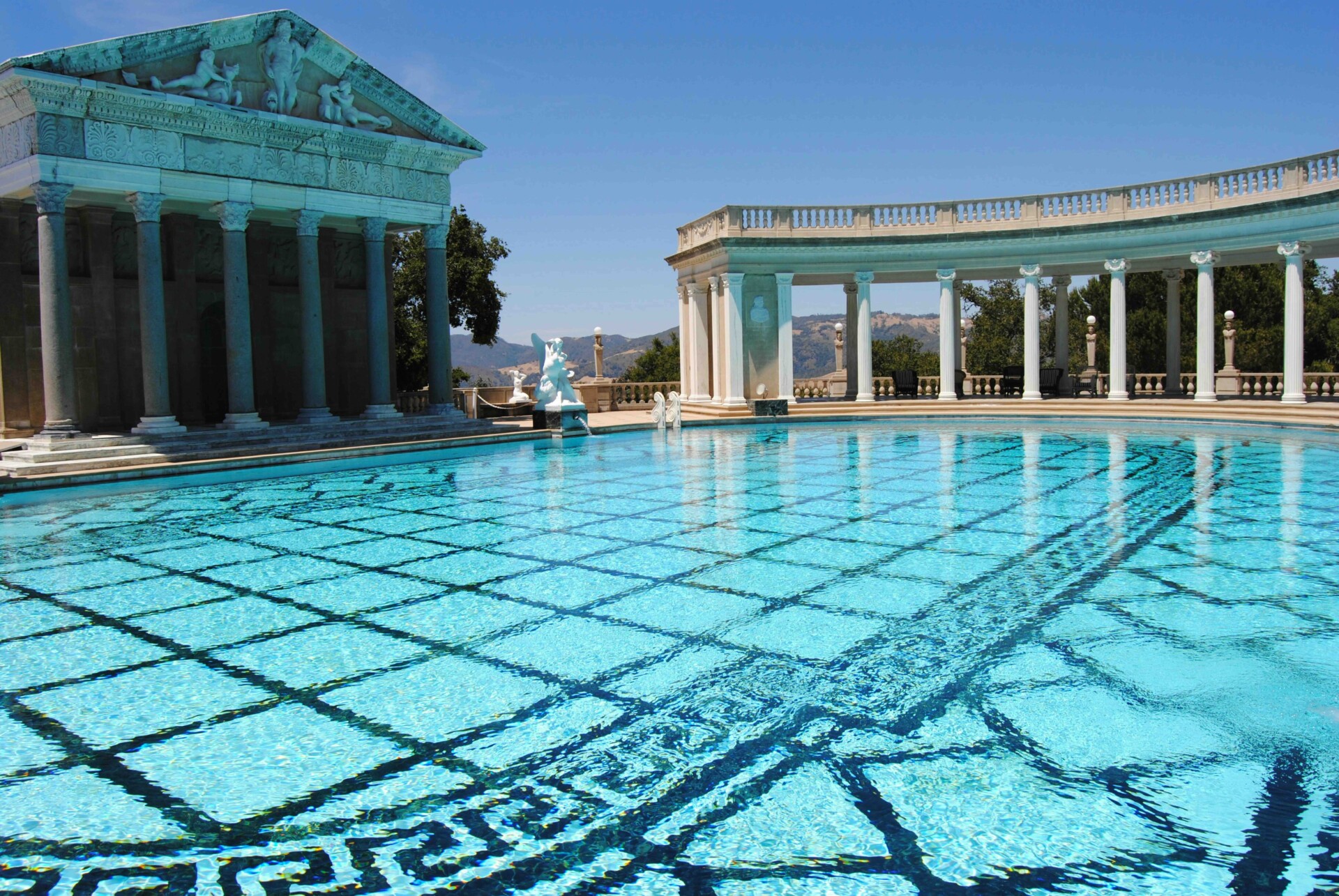 Grand outdoor pool with clear water, with Roman-like architecture surrounding the outside of the pool. Pillars positioned just outside, Hearst Castle