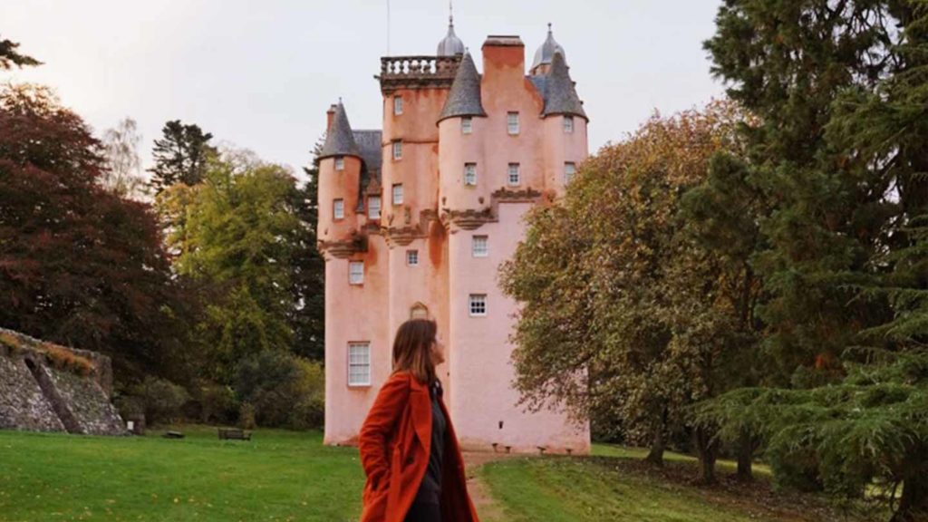 Woman in red coat looking towards a pink castle surrounded by green trees.