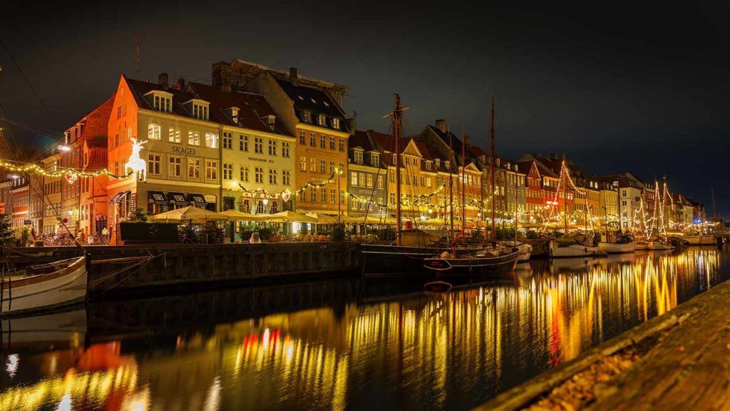 Evening view of historical European waterfront, one of the most Christmassy destinations in the world, with illuminated buildings and moored sailboats.