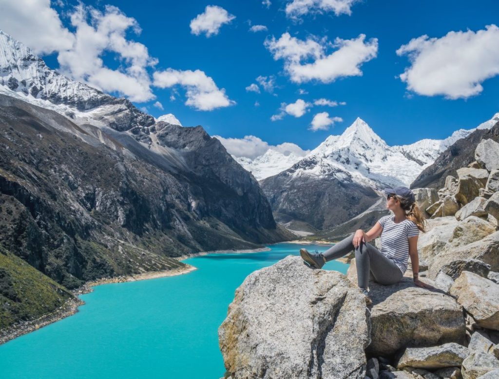 A traveler sits on a rocky outcrop enjoying the view of a turquoise glacial lake and snow-capped mountains, embodying the essence of why travel solo.