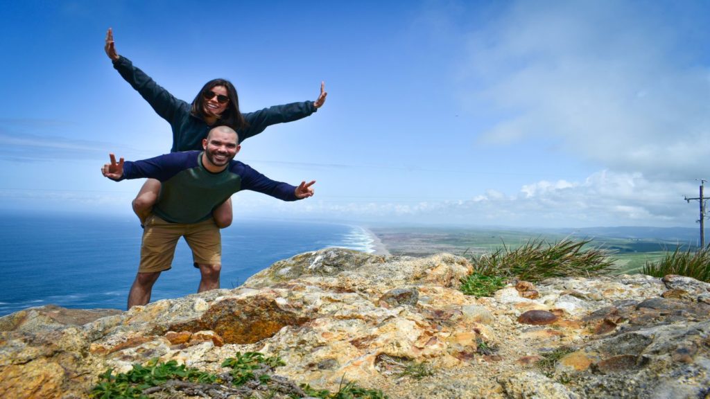 Two individuals posing with raised arms on a scenic coastal overlook, capturing one of their life-changing travel moments.
