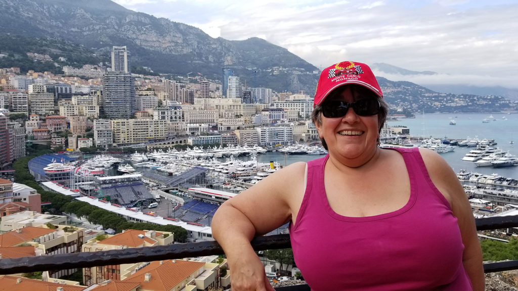 Woman in a red cap happily smiling in front of a scenic harbor filled with yachts, embodying solo travel, with buildings and hills in the background.