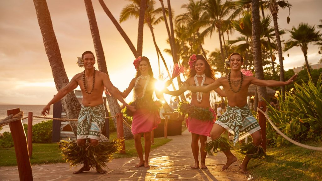 Four performers in traditional Hawaiian attire walking hand-in-hand at a tropical resort, with palm trees and a sunset in the background, celebrating Hawaiian Christmas traditions.