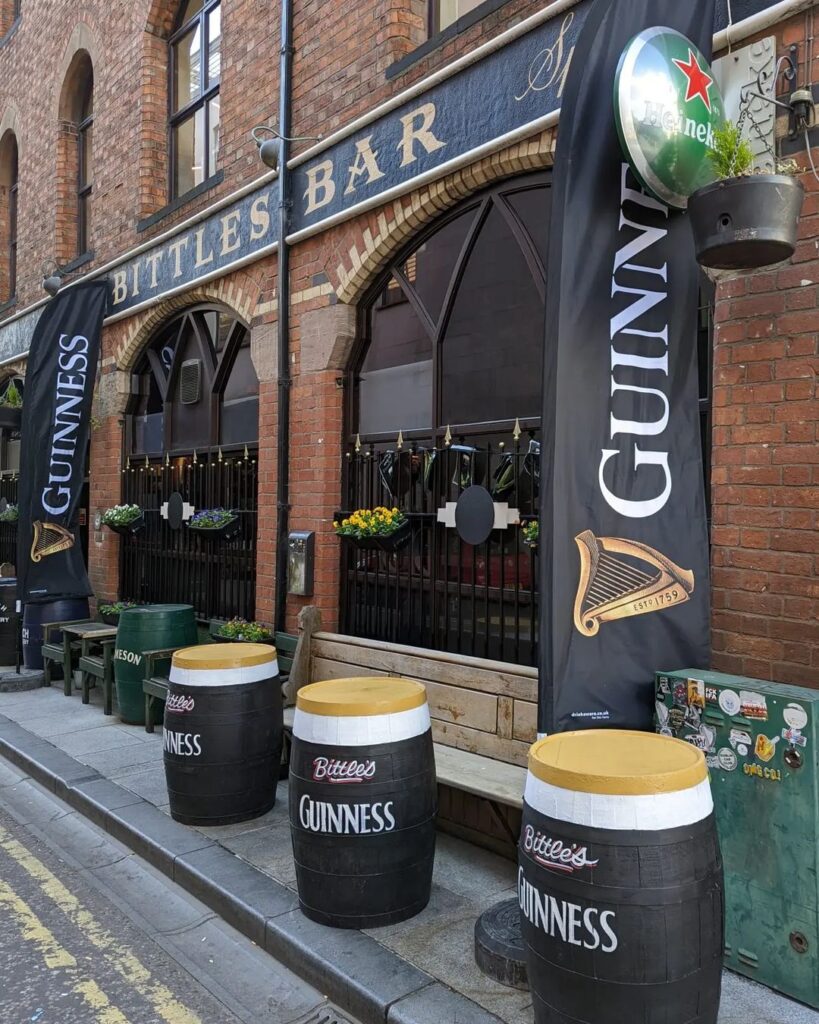 Exterior view of bittles bar with guinness branded banners and barrel tables set up on a cobblestone street, a must-visit on your European summer bucket list.