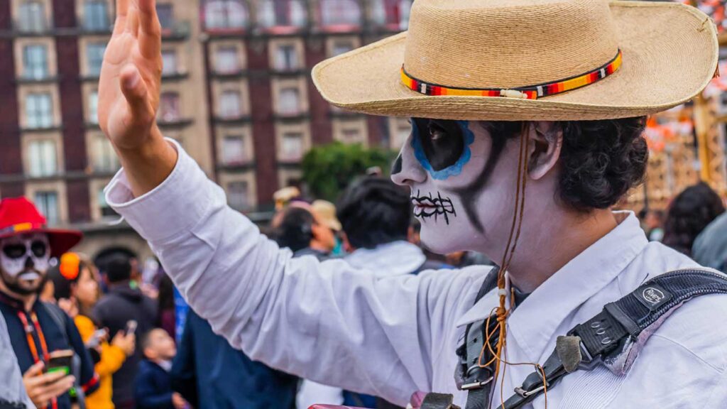 Person with skull makeup and straw hat raising hand in a crowd at a bucket list Day of the Dead celebration.