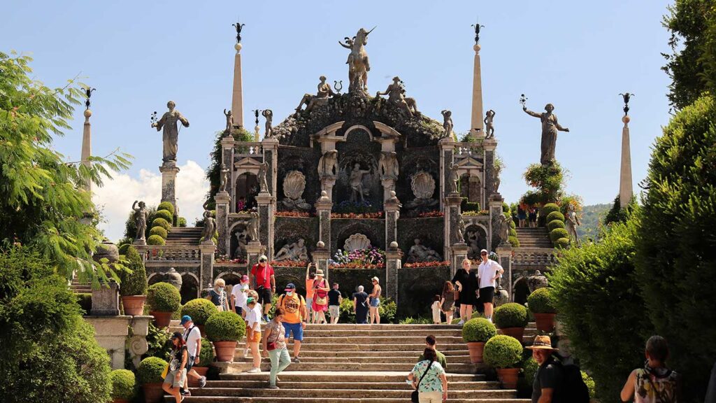 Tourists visit a grand, ornate staircase and garden with statues and lush greenery under a clear sky, often sharing hilarious travel stories.