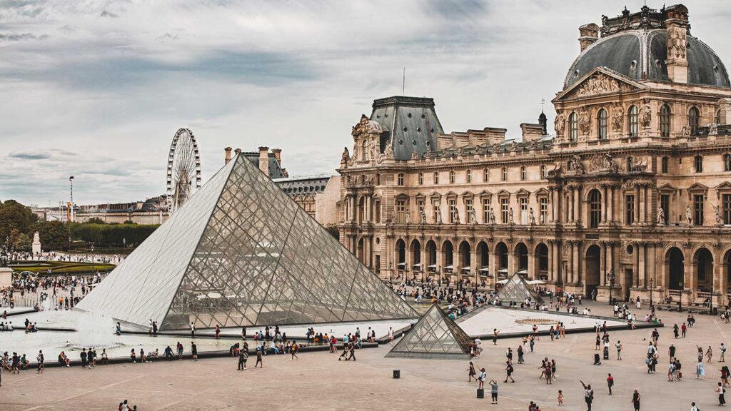 View of the Louvre Museum's glass pyramid and surrounding courtyard with visitors sharing hilarious travel stories, set against the historic palace and a cloudy sky.