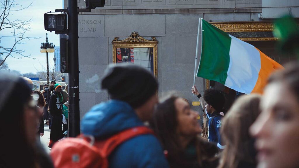A person holding an Irish flag in a bustling street scene, with people and traffic lights in the foreground during a bucket list event.