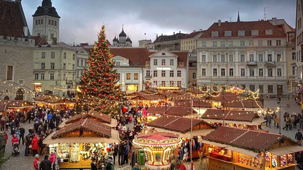 A bustling christmas market in a town square at dusk, with illuminated stalls, a decorated tree, and historical buildings under a cloudy sky is one of the bucket list events for many travelers.