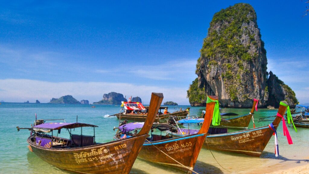 Traditional thai longtail boats anchored near a beach with a large limestone karst in the background.