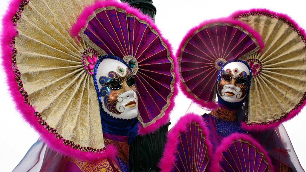 Two people in ornate Venetian masks and costumes with large, fan-like collars in purple and gold, embody a scene from Italy's annual Carnival.